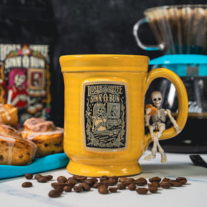 The front of the Bones Coffee Company Sinn-O-Bun hand thrown mug with the Sinn-O-Bun coffee art on the golden medallion. The mug is yellow colored. There are coffee beans and a tray of cinnamon rolls nearby.