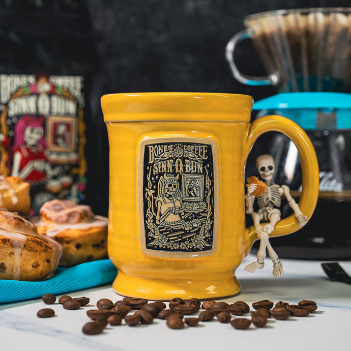 The front of the Bones Coffee Company Sinn-O-Bun hand thrown mug with the Sinn-O-Bun coffee art on the golden medallion. The mug is yellow colored. There are coffee beans and a tray of cinnamon rolls nearby.
