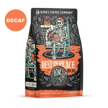 The front of a 12 ounce bag of Bones Coffee Company Rest In Peace coffee. There is a skeleton wearing a nightcap sitting on a rocking chair, reading a book and drinking coffee on the art. A sticker says decaf.