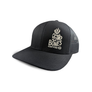The front of the skull Bones Coffee Company logo trucker hat. The hat is black and the logo has the words Bones Coffee Co with a skull above it that looks like a mug with steam coming out of the top of it.