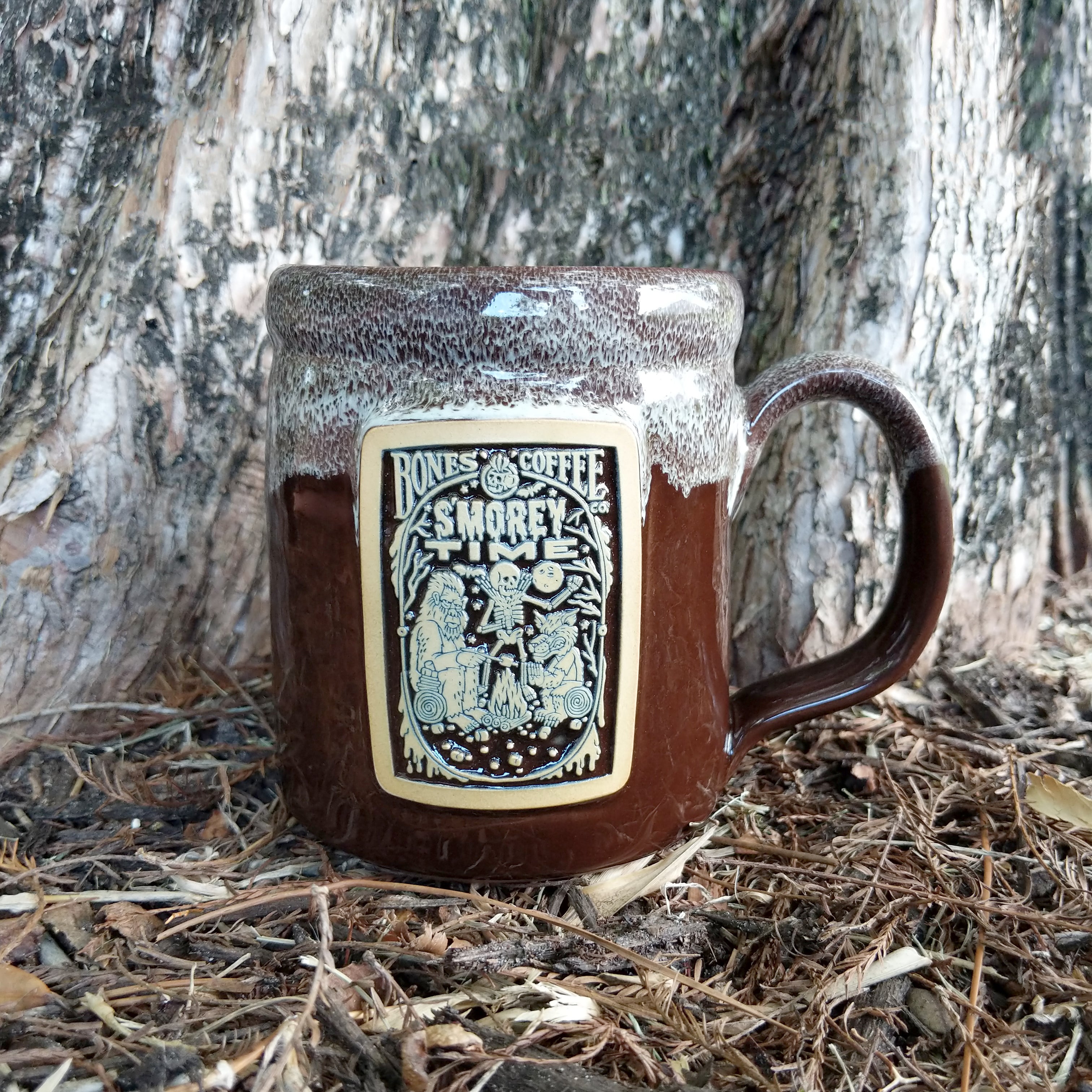 The front of the Bones Coffee Company S’morey Time hand thrown mug with the S’morey Time coffee art on the golden medallion. The mug is chocolate colored and has a white glaze on top of it.